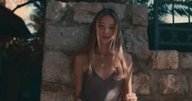 Bir kadın duvarın önünde duruyor. Siyah bir elbise ve kolye giyiyor.