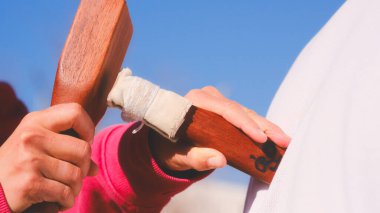 Bir kişi tahta bir çekiç ve bir sopa tutuyor. Çekiç Tayland masajında kullanılıyor. Kırmızı bir gömlek giyiyor.