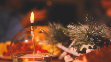 Masadaki cam kasede bir mum yakılır. Mum çam iğneleri ve böğürtlenlerle çevrili.