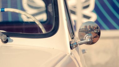 Krom aynalı ve mavi arka planı olan klasik bir araba. Ayna arabayı ve arka planı yansıtıyor, derinlik ve boyut hissi yaratıyor. Araba bir binanın önünde park halinde.