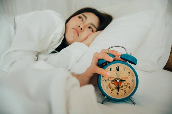Asian beautiful woman in white pajamas turning off alarm clock while sleepy in bed at her bedroom of house on holidays. Girl is having trouble wake up late in morning. Unhealthy sleep. Selective focus