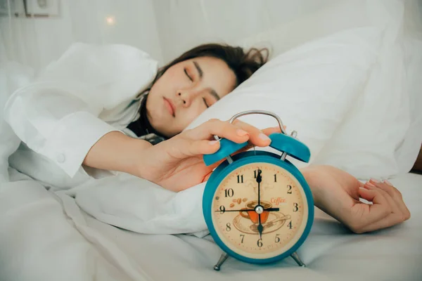 Asian beautiful woman in white pajamas turning off alarm clock while sleepy in bed at her bedroom of house on holidays. Girl is having trouble wake up late in morning. Unhealthy sleep. Selective focus