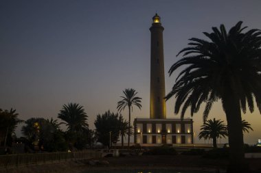 Faro de Maspalomas, Maspalomas Deniz feneri Gran Canaria 'da alacakaranlıkta ya da şafakta. Aydınlatılmış deniz feneri Suda ve palmiye ağaçlarında yansımalar.