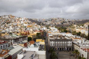 Gran Canaria, Las Palmas 'daki şehir manzarası. Mimari tarzları olan bir tepedeki binalar. Hava bulutlu. Palmiye ağaçları tropikal bir dokunuş ekler.