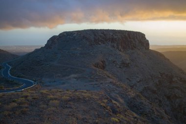 Gran Canaria 'daki dağlık bir arazide dolambaçlı bir yol. Parçalı bulutlu gökyüzü ile dramatik ışıklandırma. Mirador Astronomico de la Degollada de las Yeguas