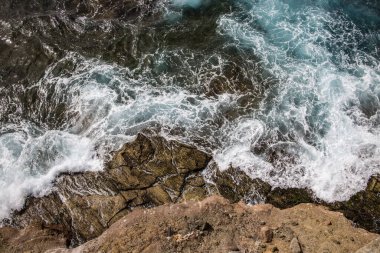 Kıyı şeridinde kayalara çarpan dalgaların hava görüntüsü. Su ve kayalardaki dinamik desenler ve dokular. Güçlü ve evcilleştirilmemiş doğa sahnesi.