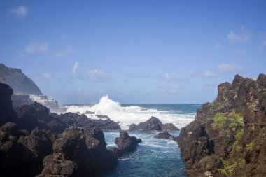 Ziyaretçiler Madeiras Piscinas Naturais do Aquario 'nun büyüleyici doğal güzelliğinin tadını çıkarıyorlar. Dalgalar parlak mavi gökyüzü altında kayalık kıyılara çarpıyor ve muhteşem bir manzara yaratıyor..