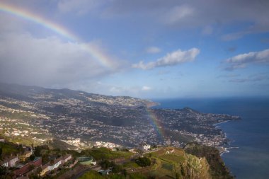 Ziyaretçiler Cabo Girao Skywalk 'ta durup Madeira' nın kıyı şeridini, yemyeşil tepeleri ve yağmurdan sonra gökyüzünde yükselen canlı gökkuşağını seyrediyorlar..