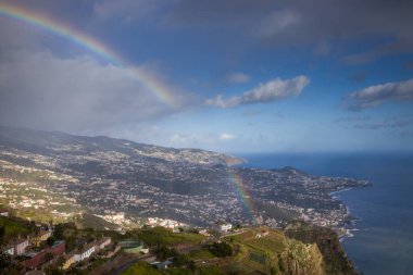 Ziyaretçiler Cabo Girao Skywalk 'ta Madeira' nın kıyı şeridi ve bereketli manzaralarının panoramik manzarasına hayret ederek duruyorlar. Canlı bir gökkuşağı manzaralı güzelliği güçlendirir ve büyülü bir atmosfer yaratır..