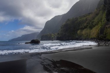 Dalgalar, Madeira 'daki Seixal Liman Sahili' ndeki siyah kuma nazikçe çarparak, yüksek uçurumların manzarayı çerçevelemesini sağlar. Rahatlamak ve doğayı takdir etmek için sakin bir yer..