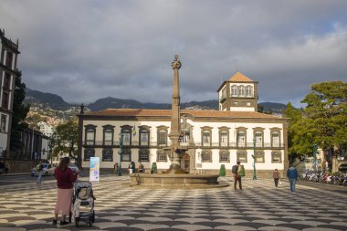 Ziyaretçiler Funchal, Madeira 'da canlı bir meydanda geziniyorlar. Tarihi mimari, merkezi bir çeşme ve palmiye ağaçları eğlence ve keşif için hoş bir atmosfer yaratır..