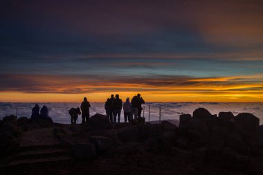 Ziyaretçiler Pico do Arieiro 'da canlı renklerle bulutların üzerinde gökyüzünü aydınlatan çarpıcı bir günbatımına tanıklık etmek için toplandılar. Hava sakin ve alacakaranlıkta manzaralı..