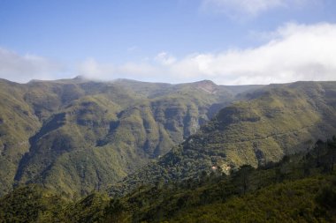 Madeira 'nın görkemli dağları canlı mavi bir gökyüzünün altında yükseliyor, yamaçları kaplayan yeşillikler ve yukarıdaki bulutların sıçraması, huzurlu doğal bir manzara yaratıyor..
