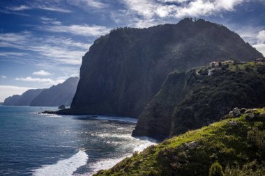 Bereketli yeşillik, derin mavi okyanusla güzel tezat oluşturan Madeira kayalıklarını kaplar. Dalgalar, berrak bir gökyüzünün altındaki kayalara hafifçe çarparak huzurlu bir sahil atmosferi oluşturur..