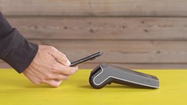 Modern kablosuz ödeme bankacılığı POS terminali, ahşap arka planda sarı masadaki dokunmatik ekranlı. NFC teknolojisiyle telefonla ulaşılamaz ödeme kavramı. Yakın plan, kenar görünüm, boşluk kopyala
