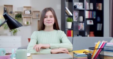 Oturma odasında masasında oturan ve kameraya gülümseyen beyaz bir genç kızın portresi. İçeride. Evde ödev yaparken güzel bir liseli kız. Zeki genç kız..