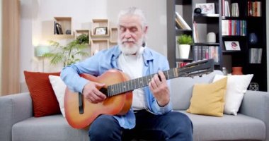 Oturma odasında koltukta oturan gri sakallı beyaz erkek besteci gitarla beste yapıyor. Yaşlı, kır saçlı adam evde müzik aleti çalıyor. Büyükbaba müzisyen..