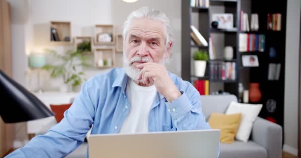 ノートパソコンを持って家に座っている間 カメラを見て思慮深刻な顔を持つ白人の古い白髪の男の肖像画 男性シニア作家とともに賢明な目でリビングルーム — ストック動画