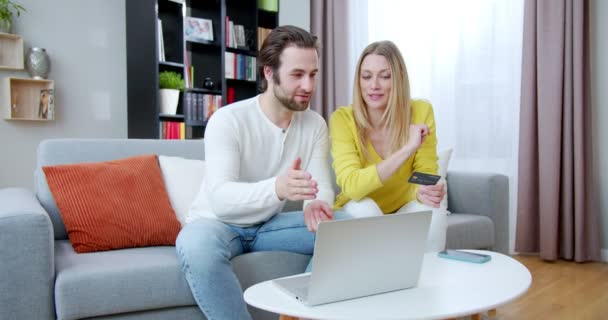 幸せな陽気な若い白人夫婦が自宅でソファに座って ラップトップコンピュータ上でオンラインショッピングをして結婚しました 男性と女性はクレジットカードで支払い インターネットで購入する — ストック動画
