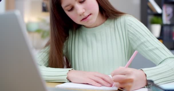 接近漂亮的白人少女坐在笔记本电脑旁 在抄袭本上写作 并在网上上课 通过互联网上学 在家里带着作业在电脑上视频聊天课的青少年 — 图库视频影像