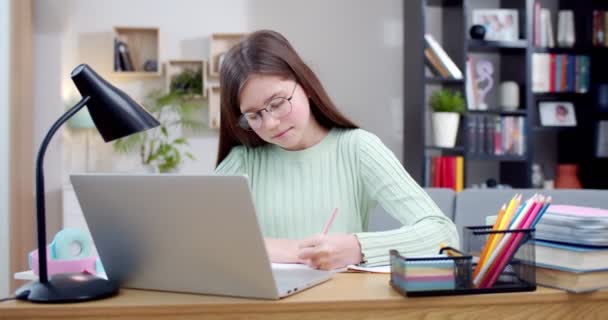 戴眼镜的好女学生坐在笔记本电脑旁 在抄袭本上写练习 并在网上上课 在网上上学 在家里用电脑上电视聊天课和写作业的青少年 — 图库视频影像