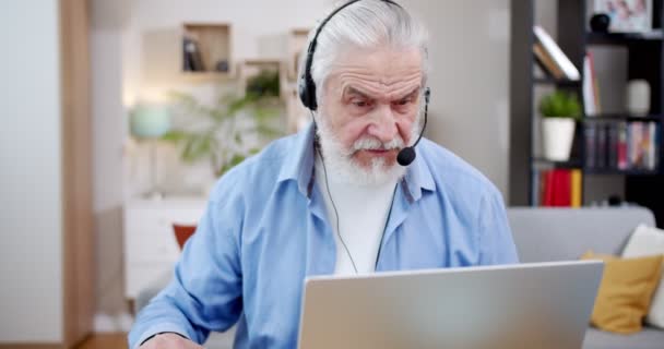 ノートパソコンのウェブカメラを介してオンラインで話をし フリーランスとして自宅で働くヘッドフォンで白人白髪の男 老人は何かを説明し リビングルームでコンピュータにビデオ通話をしています — ストック動画
