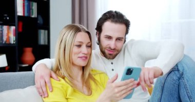 Oturma odasındaki kanepede oturan ve cep telefonu kullanan mutlu, genç, beyaz çifti kapatın. Kapalı alanda. Güzel kadın ve yakışıklı adam evde sarılıyor ve cep telefonuyla kayıyor..