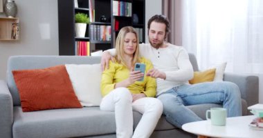 Kafkasyalı mutlu çift, akıllı telefon kullanıp bir şeyler izlerken kanepede gülüyor ve sarılıyor. İçeri gir. Neşeli erkek ve kadın cep telefonu ve kaydırma ya da mesaj kullanıyor.