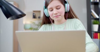 Kulaklıklı, laptopla oynayan ve evde müzik dinleyen neşeli beyaz kız. Güzel, mutlu bir çocuk rahat oturma odasında bilgisayarda sohbet ediyor ve şarkının tadını çıkarıyor. Kapalı.