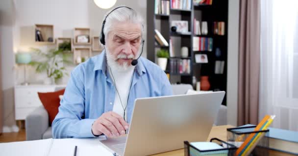 無料の仕事のための家で働くとしてノートパソコンのウェブカメラを介してオンラインで話すヘッドフォンの白人の古い白髪の男 居心地の良いリビングルームでコンピュータ上でビデオチャットを持っているシニア男性フリーランス — ストック動画