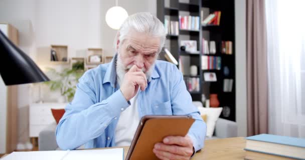 白种人的白发苍苍的祖父坐在客厅里 用平板电脑 老年人 头发灰白 看视频 在电脑上打滚 家中携带小玩意的退休男性 — 图库视频影像