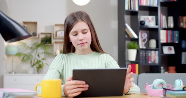 可爱的白人少女坐在舒适的客厅的桌子旁 一边笑一边看平板电脑上有趣的视频 在家里漂亮而快乐的少女在电脑上打拍子和打滚 — 图库视频影像