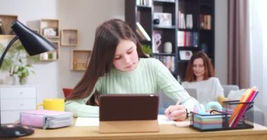 Kızım tabletle çalışıyor. Konsantre çocuk ev ödevlerini online teknoloji kitabı yakınlaştırma parşömeni ile yapıyor. Özgeçmişinde dizüstü bilgisayarla çalışan odaklanmış bir anne. Annemle aynı odada ders çalışan bir kız. Ödev konsepti.