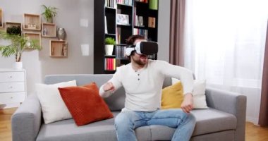 Sanal gerçeklikte video oyunu oynayan mutlu, rahat adam müzikten hoşlanıyor. VR kaskıyla dans eden güzel bir erkek evdeki kanepede oturuyor, kazak ve kot pantolon giyiyor. VR oyun konsepti.
