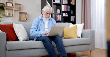 60 'lı yaşlarda laptopla evde internetten çalışıyor. Gri saçlı, yaşlı, sakallı bir adam bilgisayarda kanepeye oturmuş yazı yazıyor. Mavi gömlekli ve kot pantolonlu son sınıf öğrencisi. Teknoloji konsepti.