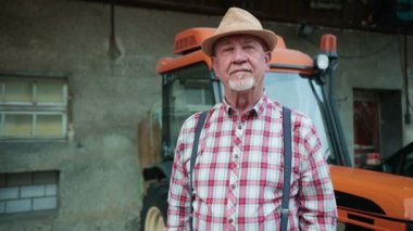 Arka planda traktörle organik çiftlik alanında duran şapkalı olgun çiftçi portresi. Adam kameraya bakıyor ve gülümsüyor.