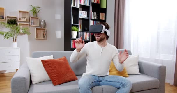 大帅哥坐在客厅的沙发上玩Vr游戏 年轻男性穿着Vr耳机 挥动着手 指尖着手 虚拟现实概念 — 图库视频影像