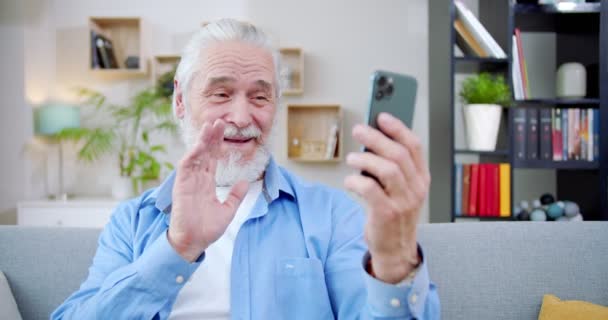 特写镜头快乐乐观的老头坐在沙发上打视频电话 容易说话的资深男性打电话给人们使用智能手机 面对面 老年人的微笑和移动的手 电话概念 — 图库视频影像