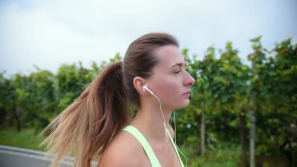 若い白人女性が屋外でイヤフォンを着て走り 音楽を聞いて閉じます 健康的なランニングと屋外運動の概念 — ストック動画