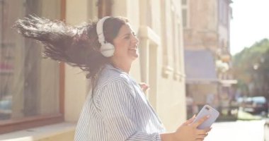Neşeli yetişkin kadın eğleniyor, akıllı telefon tutuyor ve dışarıda müzik dinliyor. Kulaklık takan, dans eden ve yazın sokakta yürüyen kadın. Eğlence, uygulama, teknoloji konsepti.