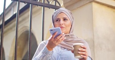 Çekici kadın Arap akıllı cep telefonu kullanıyor, dışarıda aile dostlarıyla sohbet ediyor. Genç Müslüman kadın, elinde kahve fincanı, tesettür takmış sesli mesaj gönderiyor, kaydediyor. Teknoloji, iletişim..