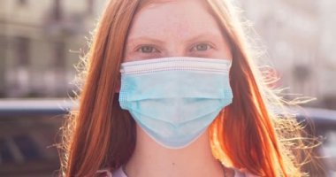 Tıbbi koruyucu maskeli, çilli güzel bir kadın sokakta duruyor. Kızıl saçlı beyaz kadın kameraya bakıyor ve güneşli bir günde gülümsüyor. Coronavirus, coviid-19, sağlık.