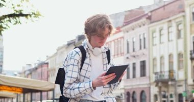 Şehirde dijital akıllı tablet kullanan sırt çantalı yakışıklı bir genç. Beyaz erkek öğrenci ekose etek giyiyor, daktilo ediyor, kaydırıyor, gadet e-postalarını kontrol ediyor. Teknoloji ve sosyal medya uyumlu.