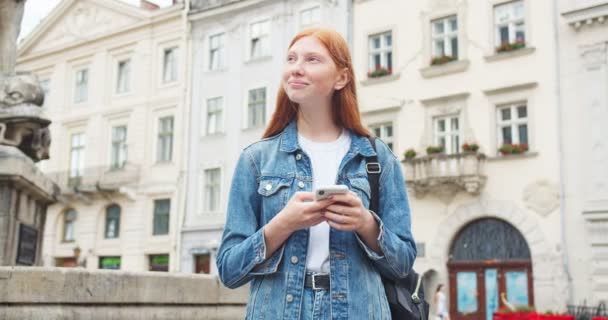 漂亮的红头发女人发短信给朋友 滚动饲料 打字的小玩意 微笑的年轻女性在智能手机上看电子邮件 在城市里到处看看 互联网通信 — 图库视频影像