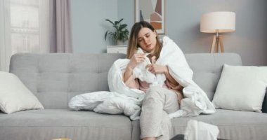 Oturma odasındaki kanepede battaniyeyle oturan beyaz bir kadın yüksek ateşi ölçüyor ve yakıyor. Grip ya da koronavirüslü sağlıksız bir kız. Ateşi, baş ağrısı ve kötü hisleri var..