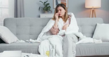 Coronavirus hastası genç beyaz kız oturma odasında kanepenin altında oturuyor, burnuna tıbbi damlalar püskürtüyor, ağlayıp hapşırıyor. Coronavirüs belirtileri gösteren hasta bir kadın..