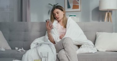 Ateşli, kanepede oturan ve akıllı telefondan video sohbeti yapan beyaz bir kadın. Hasta kızın kast etmesi ve cep telefonunda webcam ile video kaset bulundurması. Coronavirus izolasyonu.
