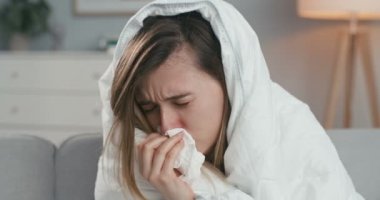 Hasta, kötü görünümlü, beyaz bir kadının evinde, kanepede oturup, hapşırması, hadım etmesi ve peçeteye burnunu silmesi. Coronavirus konsepti. Grip belirtileri gösteren bir kadın..