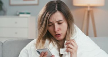 Kafkasyalı genç sağlıksız kadının battaniyeyle kanepeye oturduğu ve cep telefonuna dokunduğu ya da kaydırdığı yakın plan. Coronavirus kendini izole etme konsepti. Grip hastası bir kız akıllı telefondan mesaj yolluyor..
