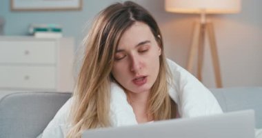 Ateşli, hasta, beyaz bir kadının evdeki kanepede oturup dizüstü bilgisayarla görüntülü sohbetini yakından çek. Hasta kız peçeteyle kast ediyor ve webcam üzerinden bilgisayar başında video kaset takıyor. Coronavirus izolasyonu.
